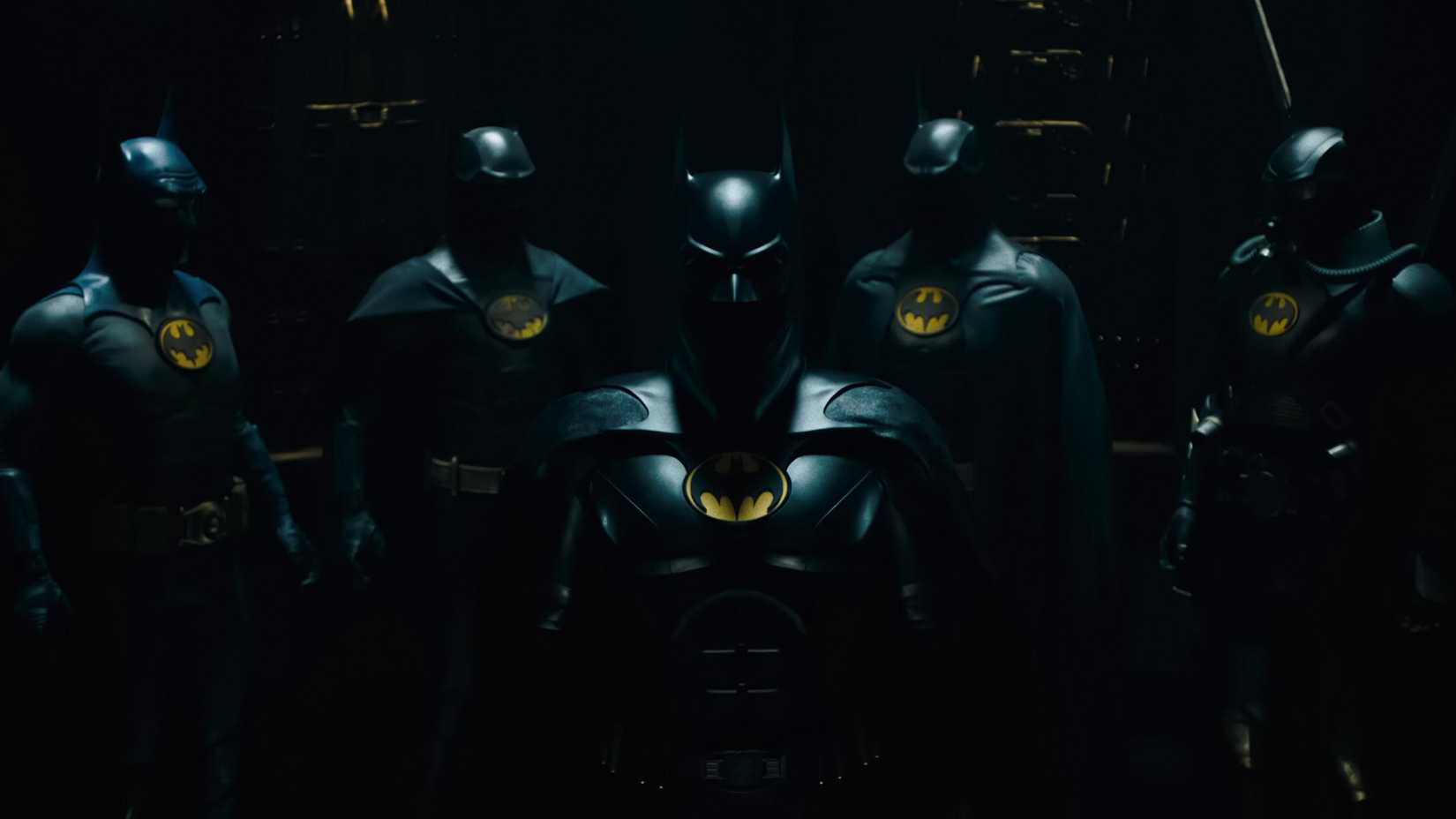 Michael Keaton as Batman with a collection of batsuits behind him from The Flash